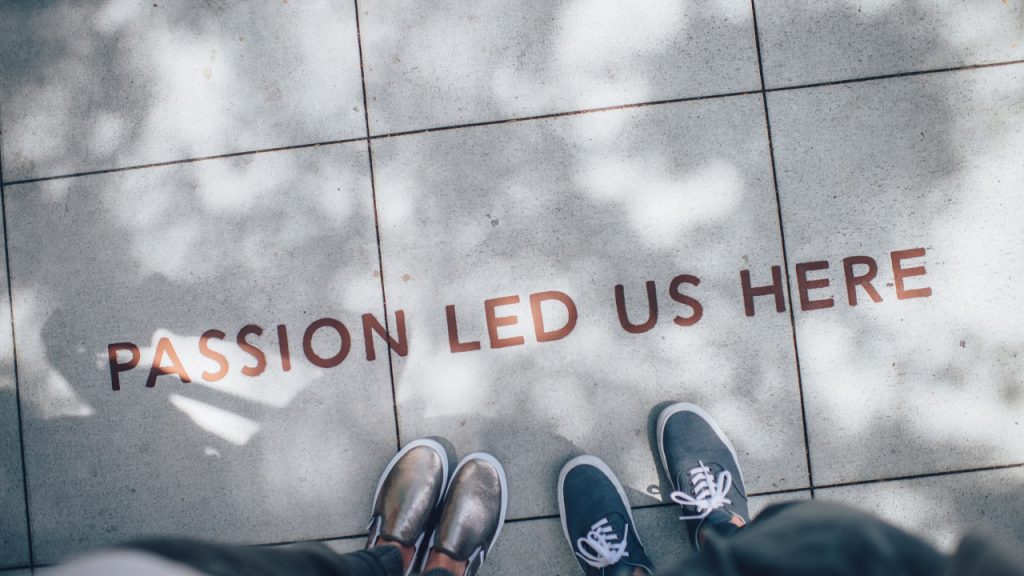 Image: Top down view of two pairs of feet walking on side walk with text sprayed onto it.
Text: PASSION LED US HERE